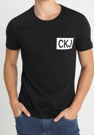 Jonge man in een zwart T-shirt met korte mouwen met "CKJ" in het wit gedrukt op de linker borst, gecombineerd met blauwe jeans.