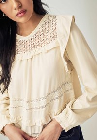 Cremefarvet bluse med blomsterløs yoke, voluminøs halsudskæring og manchetter, struktureret stof og vandret lace trim i taljen.