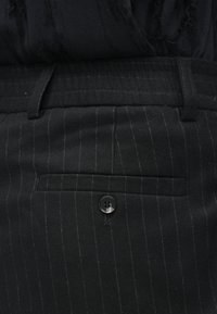 Κοντινό πλάνο μαύρων παντελονιών ρίγας με κλείσιμο τσέπης πίσω και θηλιές ζώνης, σε συνδυασμό με μια υφασμάτινη μαύρη μπλούζα.