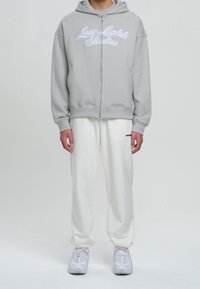 Mann trägt einen grauen Kapuzenpullover mit Reißverschluss und dem Text "Low Lights Studios", weiße Jogginghosen mit kleinem Logo und weiße Sneaker, steht vor weißem Hintergrund.