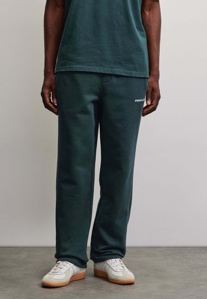 Uomo con pantaloni della tuta verde scuro e maglietta coordinata con scarpe da ginnastica bianche, in piedi davanti a uno sfondo semplice.
