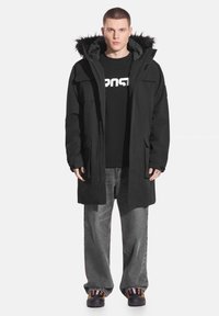 Czarna parka z futrzanym kapturem, wyposażona w wiele kieszeni i o luźnym kroju, noszona na czarnej koszulce z nadrukiem i szarych spodniach.