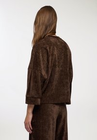 Top corto in velluto marrone con maniche ampie e vestibilità comoda. La texture appare morbida, con una lucentezza sottile e un motivo verticale.