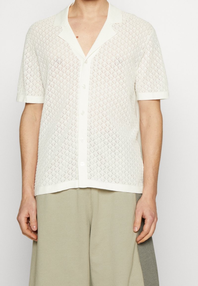 Camisa blanca de manga corta con un patrón de diamantes texturizado, cuello con muesca y botones en la parte delantera, combinada con pantalones de color claro.