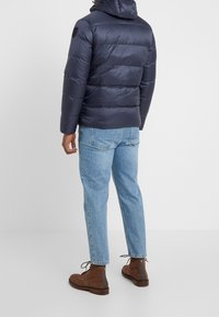 Marinblå puffjacka med huva, med horisontell vadderad design. Bärs ihop med ljusblå jeans och bruna stövlar, vilket visar en avslappnad passform.