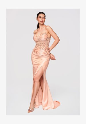 Kvinde i en gulvlængde ferskenfarvet satin kjole med detaljeret perlearbejde på livet og en slids op til låret, poserer med den ene hånd på hoften.