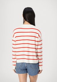 Pulover alb cu dungi orizontale roșii, cu manșete cu cât și o croială lejeră, asortat cu șorturi din denim, văzut din spate.