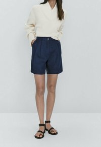 Pull à côtes de couleur crème avec col châle, associé à un short tailleur bleu marine avec un bouton et deux poches latérales. Sandales noires.