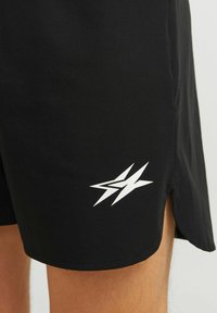 Schwarze Shorts mit einem weißen, eckigen Stern-Logo nahe dem Saum und einem kleinen seitlichen Schlitz, getragen an einem Bein.