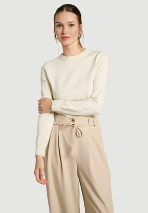 Young woman with light skin and brown hair wears a cream sweater, beige high-waist pants with tie belt, and gold hoop earrings.