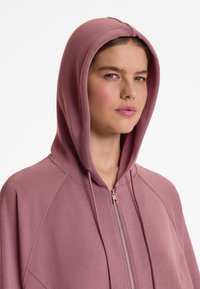 Helle rosa Kapuzenjacke aus strukturiertem Stoff, mit durchgehendem Reißverschluss, vorderer Tasche und Kapuze. Das Design umfasst Raglanärmel und einen weichen Fall.