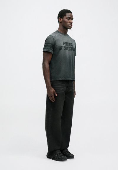 Schwarzes T-Shirt mit kurzen Ärmeln, verwaschenen Details und großem Drucktext "PSEUDO INTELLECTUAL", kombiniert mit sackartigen schwarzen Hosen und schwarzen Schuhen.