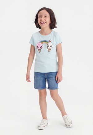 Smilende barn med kort mørkt hår iført en lyseblå skjorte med to tegneserie-isvafler og en regnbue, cowboyshorts og hvide sneakers.