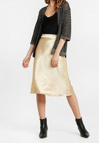 Satin guldkjol med A-linjeform, tillsammans med en svart, draperad, ärmlös topp och en texturerad svart kofta. Svarta ankelstövlar.