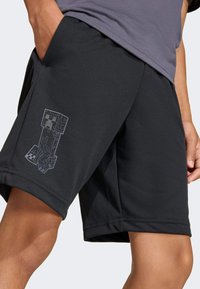 Crne sportske kratke hlače s glatkom teksturom i elastičnim pojasom. Imaju sivi pikselski grafički uzorak na lijevoj nozi. Prozračan materijal.