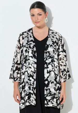 Donna con capelli scuri raccolti in uno chignon elegante, indossa orecchini a cerchio bianchi dalla texture evidente e un kimono semitrasparente con motivo bianco e nero sopra una maglia nera.