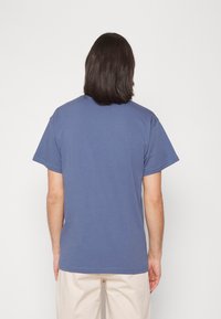 Vista posterior de una camiseta de algodón azul, de cuello redondo, mangas cortas, con un diseño sencillo y dobladillo recto, combinada con pantalones de color claro.