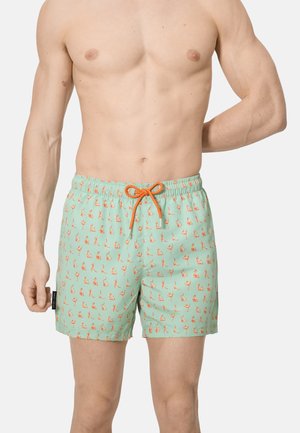 Hombre sin camiseta vistiendo pantalones cortos de baño verde claro con pequeñas ilustraciones naranjas de posturas de yoga y un cordón naranja.