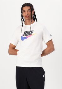 Balta medvilninė marškinėliai su dideliu juodu "Nike" logotipu, dekoruotu spalvingais gradientais, derinami su juodomis sportinėmis kelnėmis, demonstruojančiomis laisvą stilių.