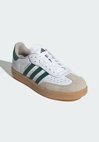 Weißer Sneaker mit grünen Streifen, Wildlederzehenkappe und durchbrochenen Details. Verfügt über eine sandfarbene Gummisohle und Schnürsenkel über einer gepolsterten Zunge.