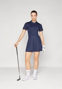 Granatowa sukienka golfowa z krótkimi rękawami, kołnierzykiem i plisowaną spódnicą. Modelka trzyma kij do golfa, ma na sobie białe skarpetki i buty. Widoczna struktura tkaniny.