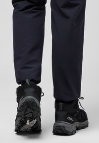 Black hiking boots with textured rubber sole, mesh upper, and reflective accents, paired with navy pants, viewed from the back.