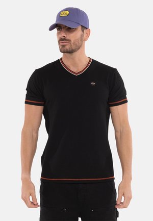 Homme portant un t-shirt noir à col en V avec bordure rouge et une casquette violette avec un logo jaune, debout devant un fond blanc uni.