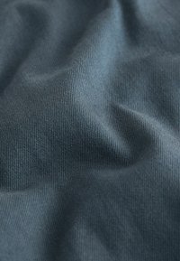 Nahaufnahme eines weichen, blau-grauen gewebten Stoffs mit sichtbarer diagonaler Textur und sanften Falten, die Schatten werfen.