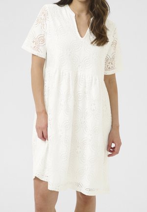 Robe blanche longueur genou avec motif en dentelle florale, manches courtes et col en V portée par une femme aux longs cheveux bruns.