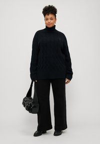 Juoda megzta turtleneck tipo liemenė su raukšlėta apačia, derinama su plačiomis juodomis kelnėmis ir juoda kryžminė rankinė. Modelis stovi prieš neutralų fono.