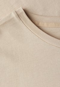 Tela de algodón beige con una textura suave, que presenta un escote redondeado y un sutil detalle acanalado a lo largo del collar.