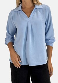 Lichtblauwe blouse met lange mouwen, een V-hals, een textuurstof en een kraag. De pasvorm is ontspannen, met een subtiele plooi in het midden.