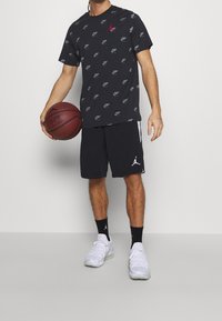 Černé tričko s opakujícím se vzorem loga, červený akcent, spárováno s černými šortkami s bočními pruhy a logem. Bílé sportovní boty. Drží basketbal.