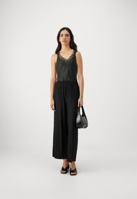 Schwarzes Satin-Camisole mit Spitzenbesatz, kombiniert mit schwarzen plissierten Hosen. Das Outfit wird mit einer schwarzen Handtasche und offenen Sandalen vervollständigt.