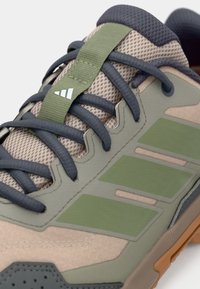 Μπεζ αθλητικό παπούτσι με πλέγμα στο πάνω μέρος, γκρι κορδόνια, πράσινες λεπτομέρειες και ελαστική σόλα με υφή. Διακριτό το λογότυπο της Adidas.