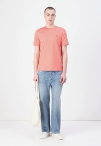 Korallfärgad bomull t-shirt med en liten gul logga, tillsammans med ljusblå raka jeans och vita sneakers. Håller en beige tote-väska.