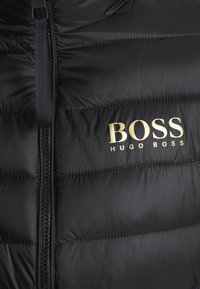Mustapuffaliivi, joka on valmistettu sileästä ja kiiltävästä materiaalista. Siinä on edessä vetoketju, tikattu muotoilu ja kullanvärinen "BOSS HUGO BOSS" -logo rinnassa.