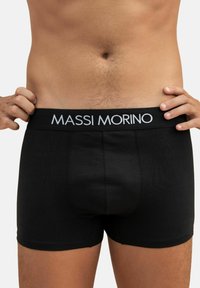 Torso maschile che indossa boxer neri con il logo "MASSI MORINO" sulla vita, mani che sistemano l'elastico.