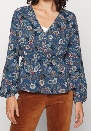 Femme portant un chemisier bleu à manches longues à fleurs avec un col en V et une taille élastique, associé à un pantalon en velours côtelé marron.