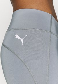 Leggings atletici grigi con una texture liscia e un logo bianco Puma sul lato. Le cuciture visibili e il design aderente conferiscono un aspetto sportivo.