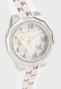 Montre octogonale argentée avec un bracelet en silicone blanc à motif. Le cadran présente un logo triangulaire doré "GUESS" et des accents en cristal.