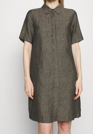 Kobieta w krótkim, do kolan, ciemnobrązowym lnianym sukienka z klasycznym kołnierzykiem i krytymi guzikami z przodu.