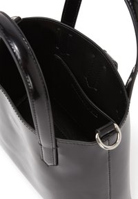 Sac fourre-tout en cuir noir avec finition brillante, ouvert sur le dessus, au design structuré, poche intérieure à glissière et accents en métal argenté. Texture lisse dans l'ensemble.