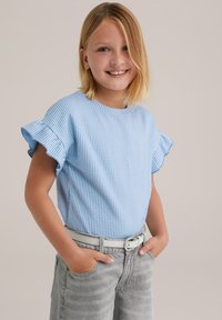 Mosolygó, szőke hajú gyermek világoskék csíkos rövid ujjú pólóban és szürke farmernadrágban, a zsebébe dugott kezekkel, sima háttér előtt.