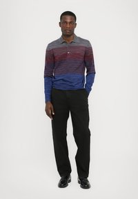 Långärmad polo-shirt i gradientränder av röd, lila och blå, med krage. Kombinerad med svarta byxor och blanka svarta skor.