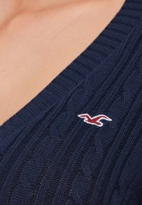 Tume sinine kudumissviiter, millel on nöörmustrit ja sügav V-kaelus. Vasakul küljel on punane ja valge tikandiga logo.