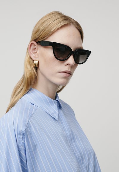 Occhiali da sole neri angolari con lenti scure, indossati con una camicia a righe blu chiaro. Un orecchino a cerchio dorato aggiunge contrasto al look.