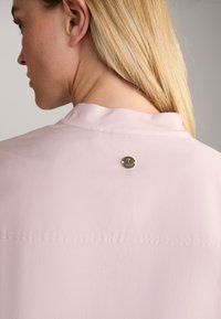 Bluse in Hellpink mit glatter Textur. Verfügt über einen Kragen, einen Knopf auf der Rückseite und dezente Stickereien an den Schultern.