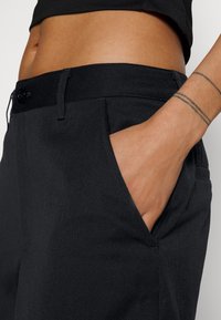 Černé zúžené kalhoty s hladkou texturou, vybavené bočními kapsami a zapínáním na knoflík. Pas je elastický pro větší pohodlí.