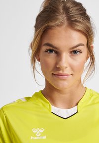 Lys gul sportstrøje med korte ærmer, sorte accenter og V-hals design. Har logo på brystet og hvide striber på skuldrene.